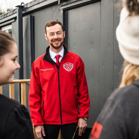 A person in a red jacket with a logo, standing in front of a grey building. 1