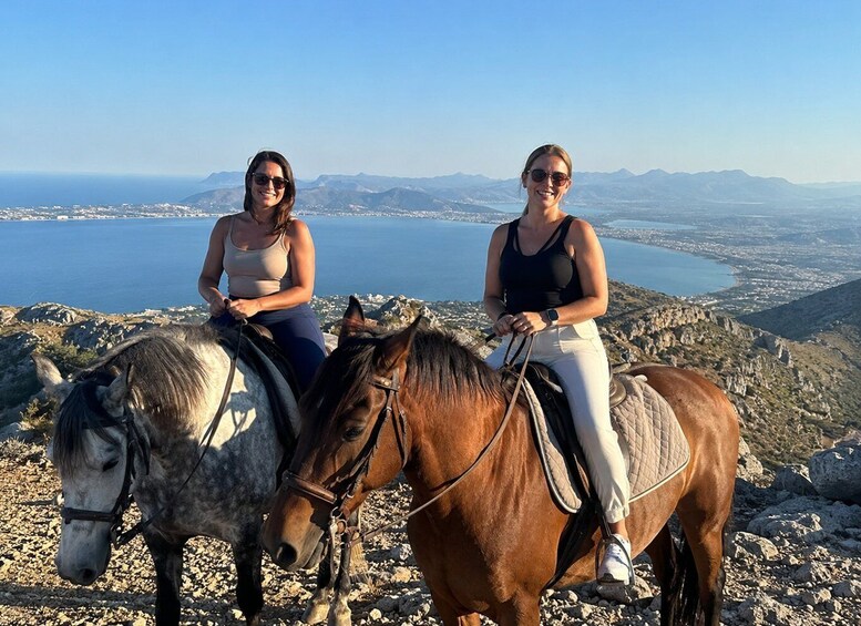 Two women riding horses on a mountain overlooking a coastal landscape. 1