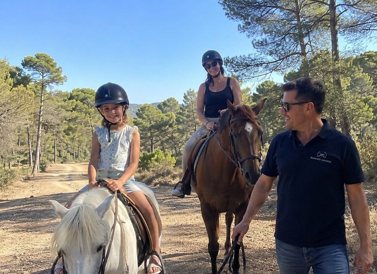 A person leading a horse and two people riding horses on a dirt path with trees in the background. 5