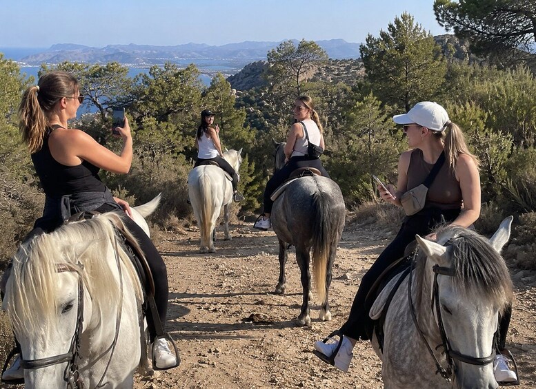 Three people horseback riding on a dirt trail with a scenic view of mountains and trees in the background. 4