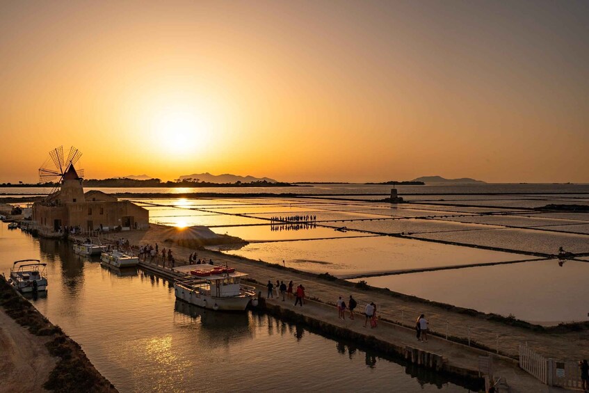 A sunset over a water body with boats, a windmill, and people walking along the shore. 2