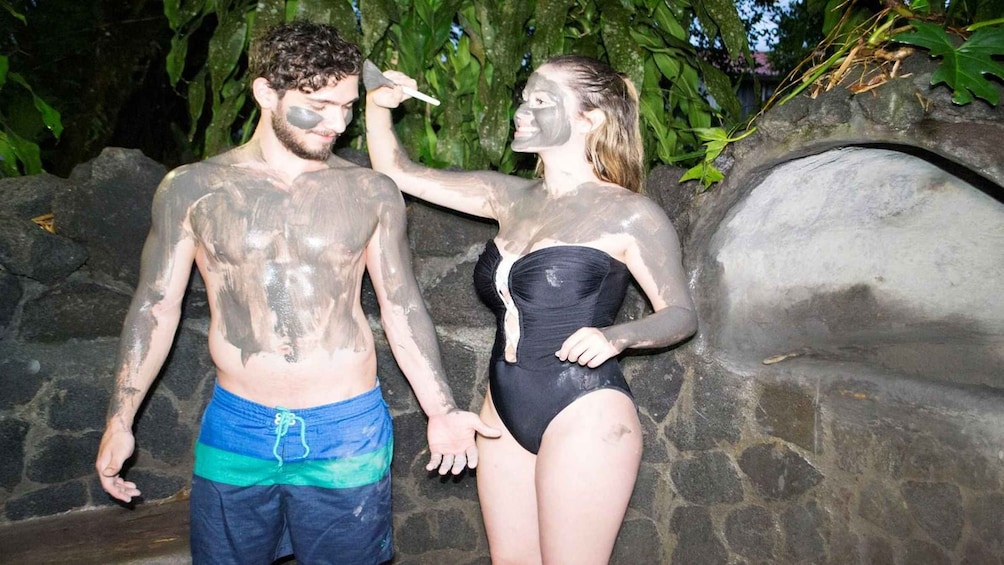 Two people covered in mud, standing in front of a stone wall with greenery in the background. 4