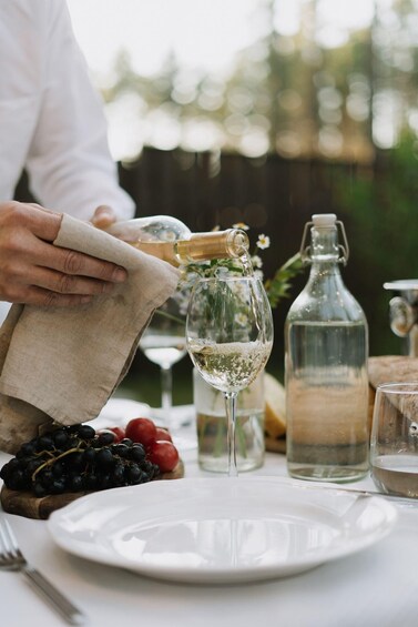 A person pouring a clear liquid from a bottle into a glass on a table with a plate, grapes, and a bottle of water. 3