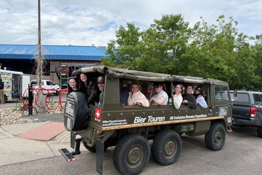 A group of people in a military-style vehicle with 'Bier Touren' branding, parked on a street with a building in the background. 1