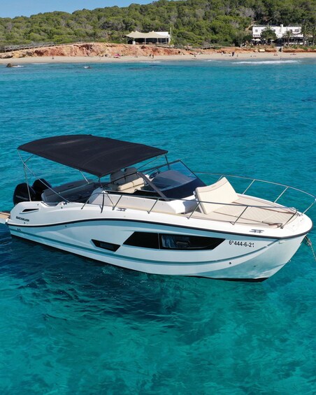 A white motorboat with a black canopy on clear turquoise water, docked near a beach with a white building and trees in the background. 5