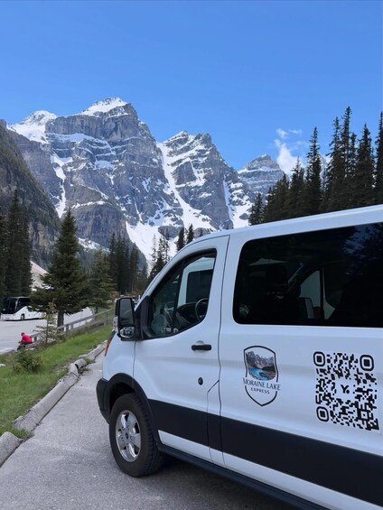 A white van parked on a road with a QR code and 'Moraine Lake Express' text, surrounded by snow-capped mountains and pine trees. 2