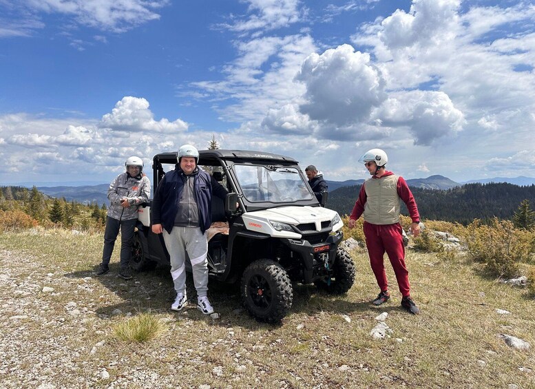Four people standing beside a white off-road vehicle on a rocky terrain with mountains in the background. 1