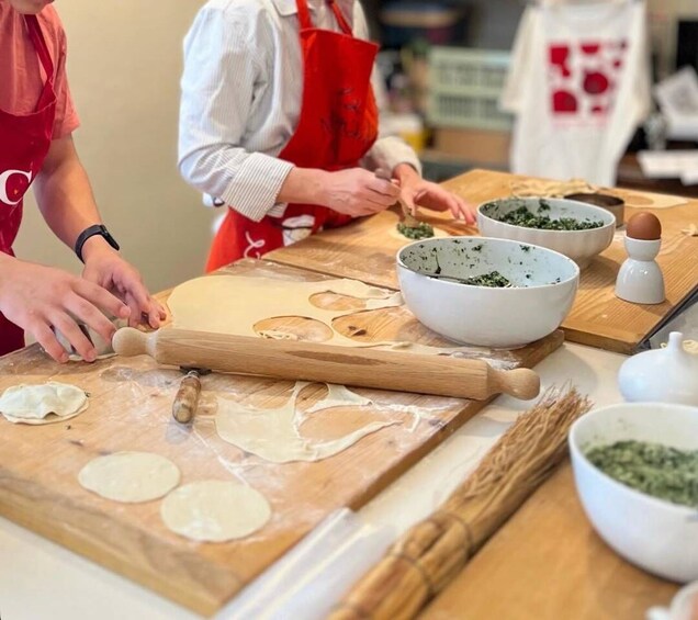 Two people rolling out dough on a wooden surface with rolling pins, bowls of green mixture, and a white container. 1
