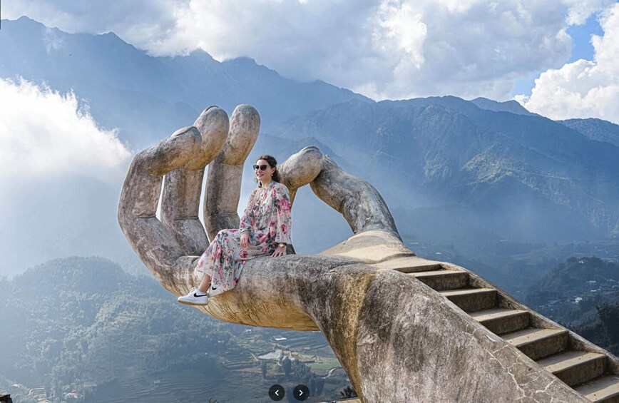 A person sitting on a large hand-shaped structure with a mountainous landscape in the background. 2