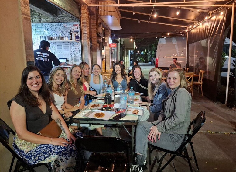 A group of people seated at an outdoor table with food and drinks, under a covered area with string lights. 4