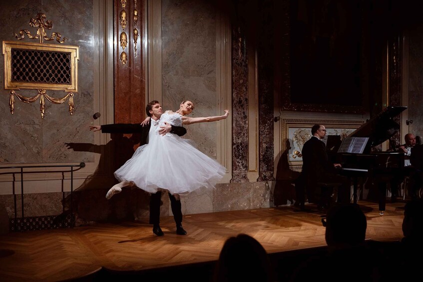 A ballet performance on a wooden stage with a grand piano and a decorative wall in the background. 3