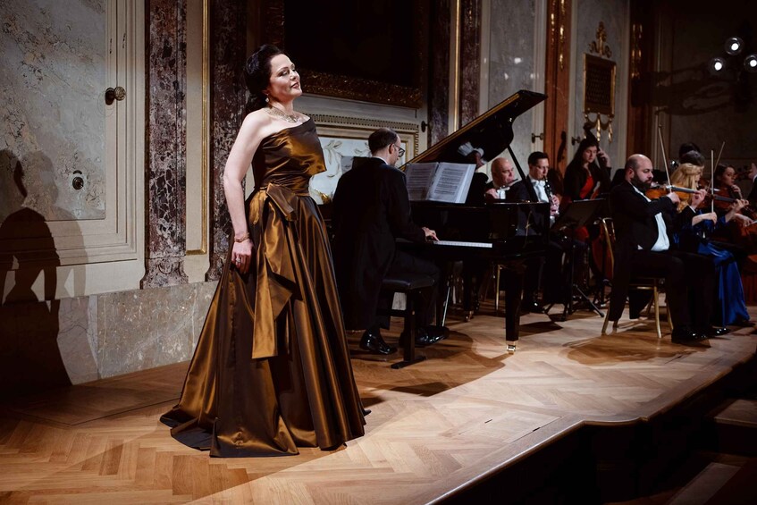 A woman in a long gown standing on a stage with a piano and orchestra. 5