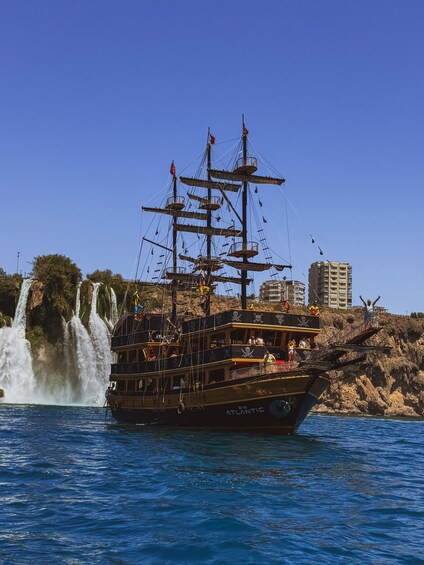 A tall ship with multiple masts and a pirate-themed design, sailing near a waterfall and rocky coastline. 5