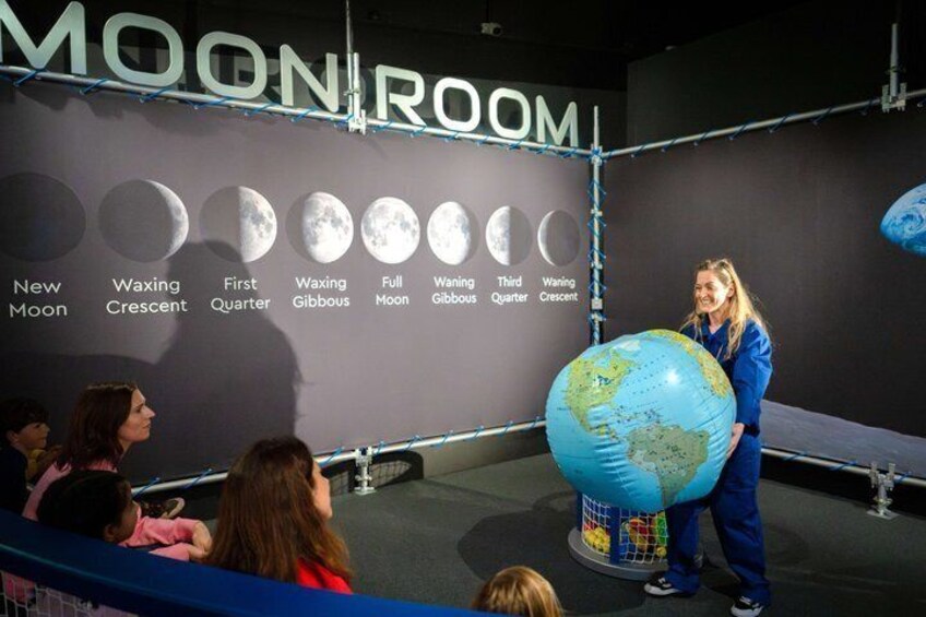 A person presenting a globe in a room labeled 'MOONROOM' with moon phases depicted on the wall. 3
