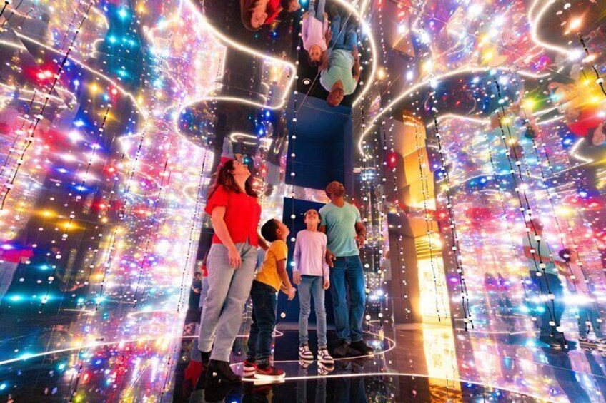 A group of people standing in a brightly lit, futuristic environment with vibrant, multicolored lights and geometric patterns. 1