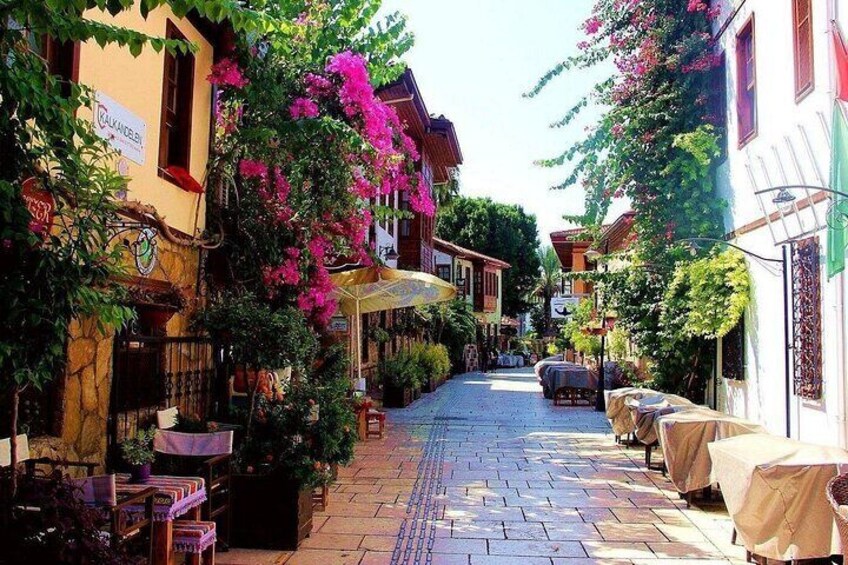 A narrow street with buildings on either side, outdoor seating, and flowering vines. 3