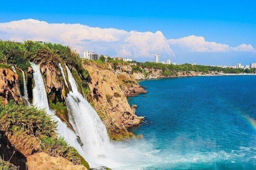 A coastal landscape with a waterfall, clear blue sea, and distant city skyline. 2