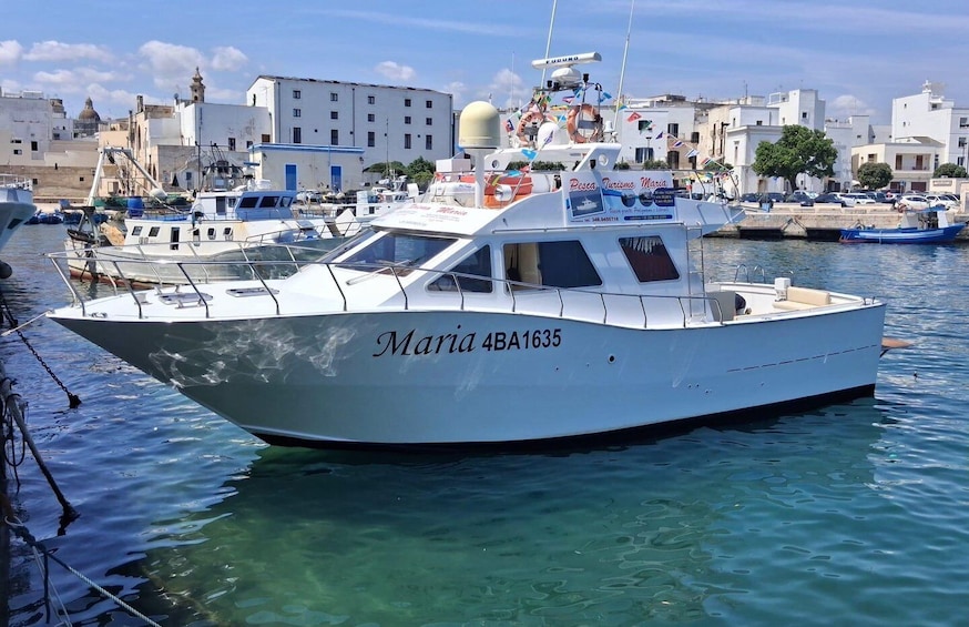 A white boat named 'Maria 4BA1635' docked at a marina with buildings and other boats in the background. 4