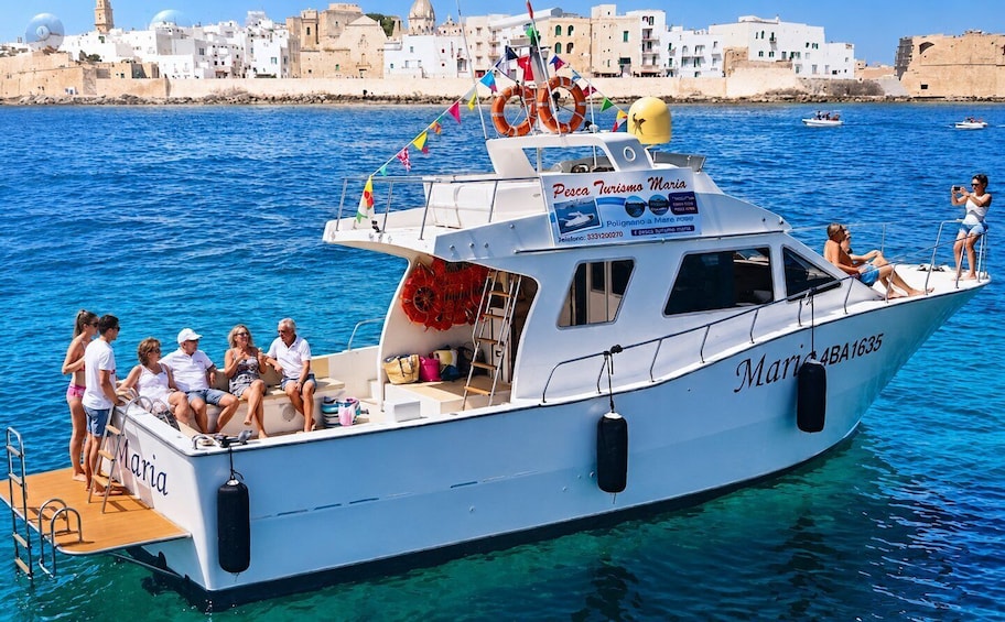 A white boat named 'Maria' with passengers and a lifebuoy on deck, docked near a coastal town with buildings in the background. 3