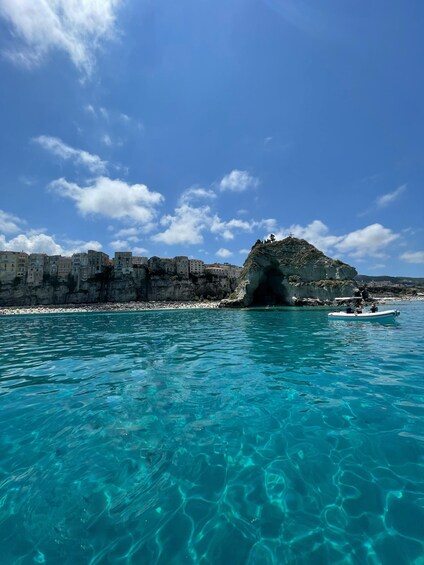 A coastal scene with clear turquoise waters, a rocky outcrop with a cave entrance, and a boat in the distance. 2