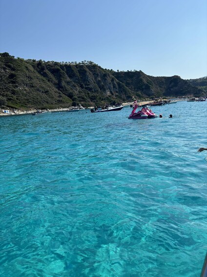 A clear blue sea with boats, a pink watercraft, and a hilly coastline. 5