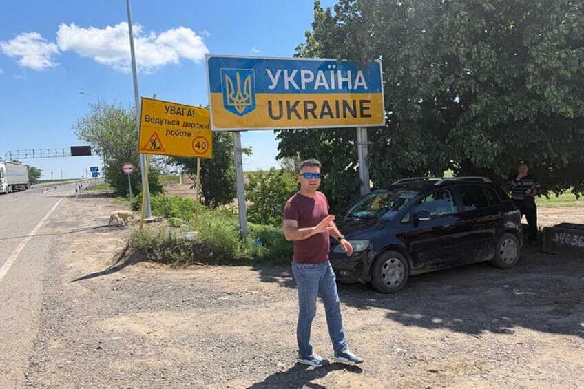A man standing next to a road with a blue sign that reads 'UKRAINA' and a yellow sign with a pedestrian symbol and the number 40. 2