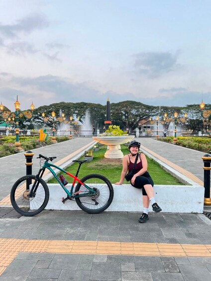 A person sitting on a bench with a bicycle beside them, in a park with a fountain and decorative lights in the background. 2