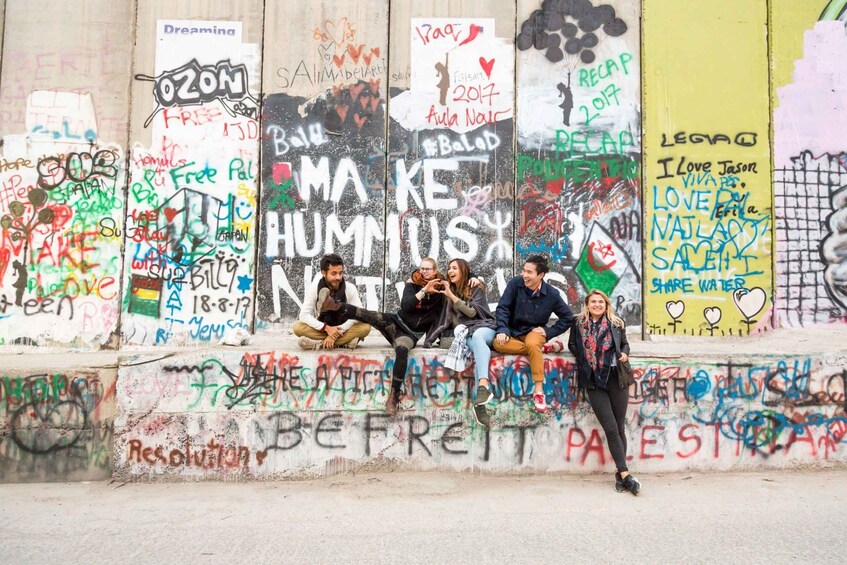 A wall covered with graffiti and messages, with people sitting and standing in front of it. 1