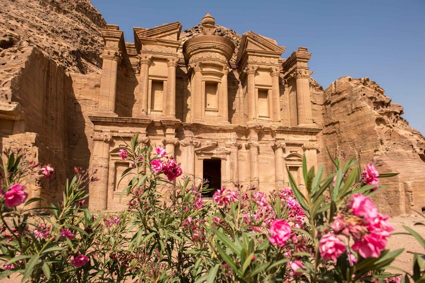 A large sandstone structure with columns and a central dome, surrounded by pink flowers. 1