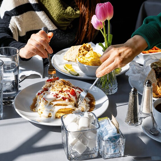 A person eating a dessert with a fork, a glass of water, and a vase with a pink flower on the table. 2
