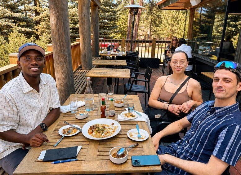 Three people dining at an outdoor wooden table with food remnants, a bottle, and a smartphone. 5