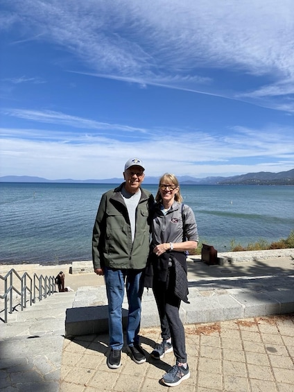 Two people standing on a stone platform by a body of water with mountains in the background. 3