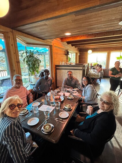 A group of people dining at a wooden table in a restaurant with large windows and potted plants. 4