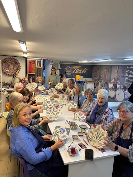 A group of people engaged in a pottery-making workshop, surrounded by pottery items and a sign reading 'Testo.' 1