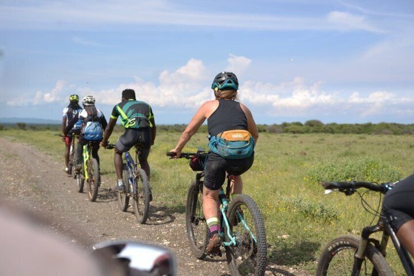 A group of cyclists on a dirt road with a grassy field and distant hills in the background. 2