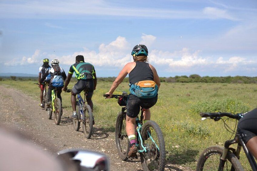 A group of cyclists on a dirt road with a grassy field and distant hills in the background. 2