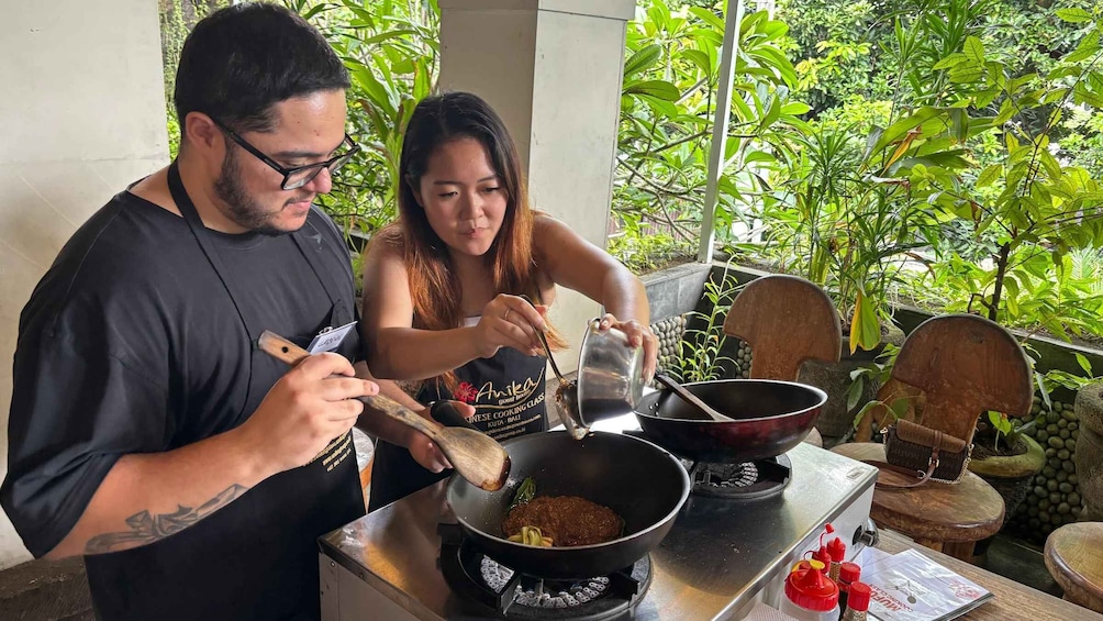 Two people cooking outdoors with a wok on a stove, surrounded by greenery. 1