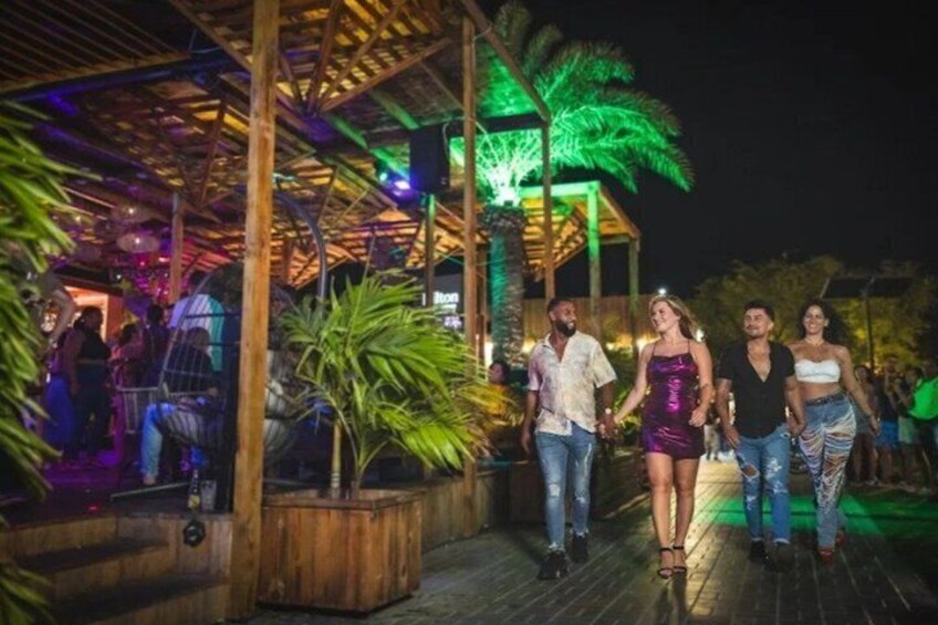 A wooden deck with people walking, palm trees, and a stage with colorful lighting. 3