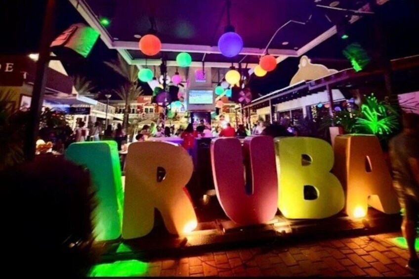 A brightly lit outdoor area with large, colorful letters spelling 'RUBA' and a crowd of people in the background. 2
