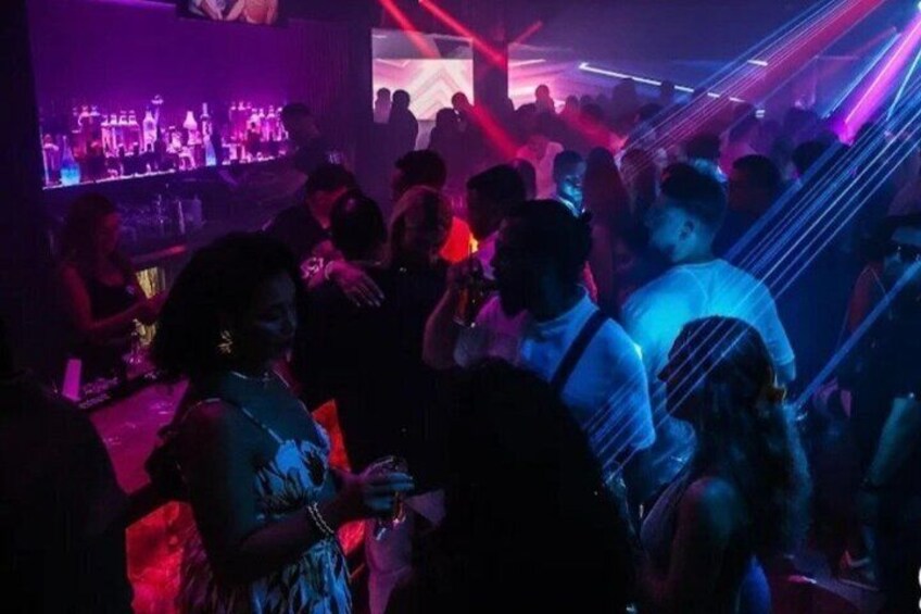 A crowded indoor social scene with people standing and conversing, illuminated by colorful stage lighting. 5