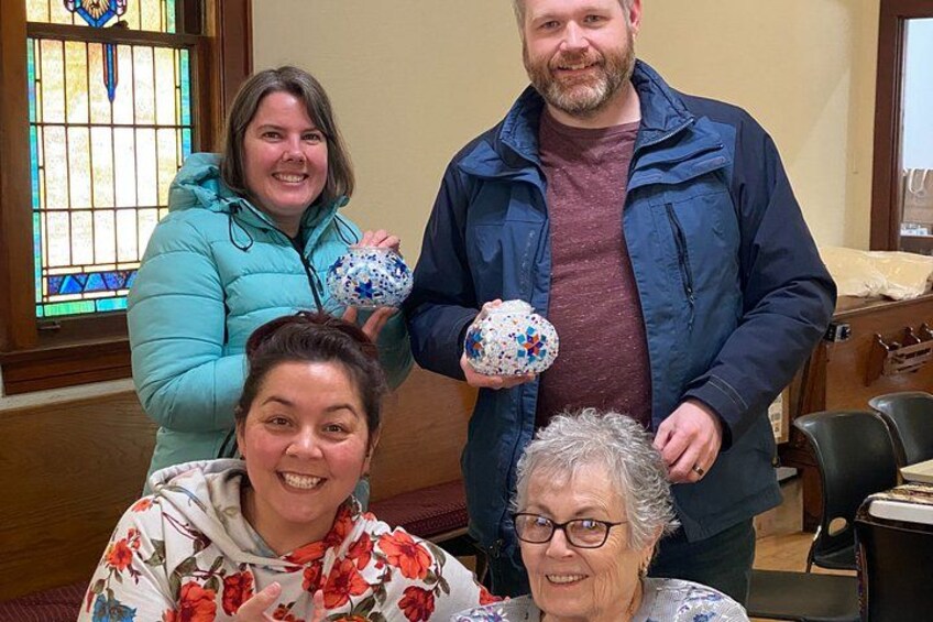 Four people indoors, two holding decorative balls, with a stained glass window and wooden furniture in the background. 5