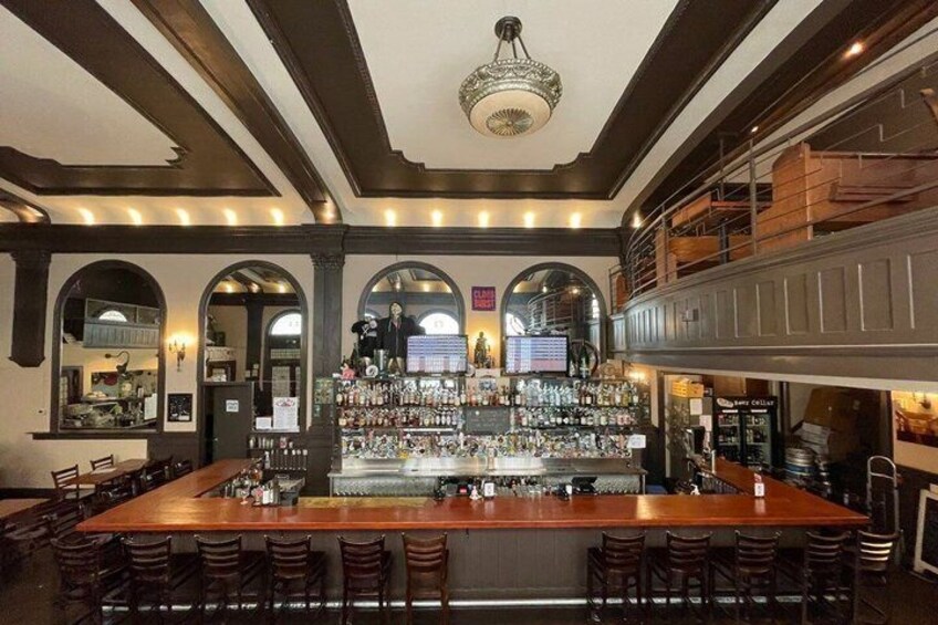 A bar with a long counter, multiple stools, and a high ceiling with exposed beams. 2
