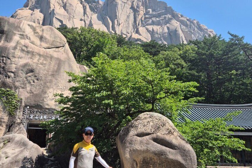 A person standing in front of a rocky mountain with trees and a traditional building. 3