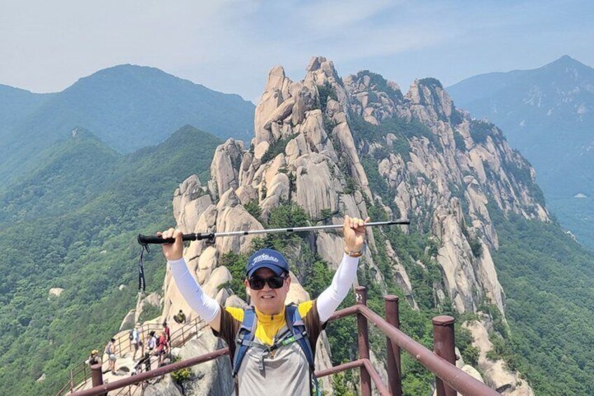 A person with a camera on a mountain trail with rocky cliffs and greenery in the background. 1