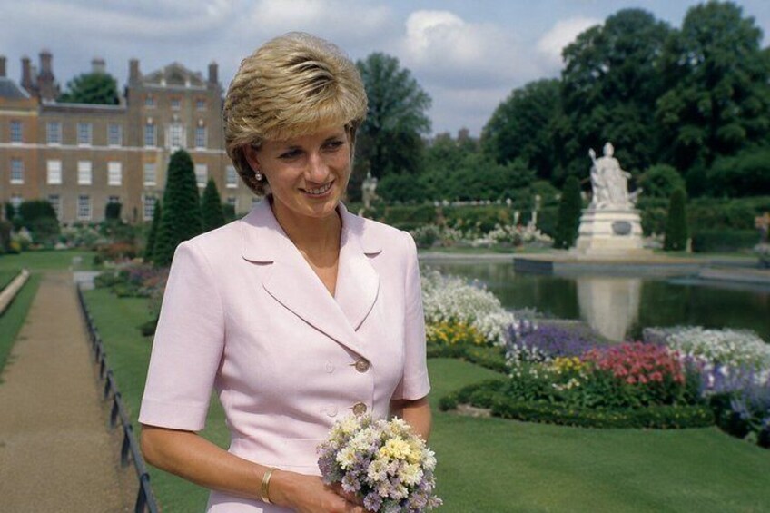 A person in a light pink suit holding a bouquet of flowers, standing in a garden with a fountain and statues in the background. 1