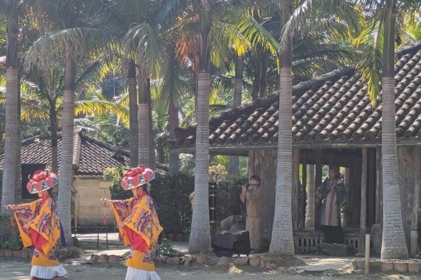 Two dancers in colorful costumes performing in front of a traditional building with a tiled roof, surrounded by palm trees. 3