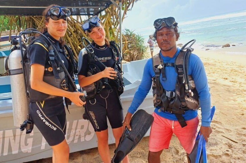 Three individuals in scuba diving gear on a beach with a boat in the background. 1