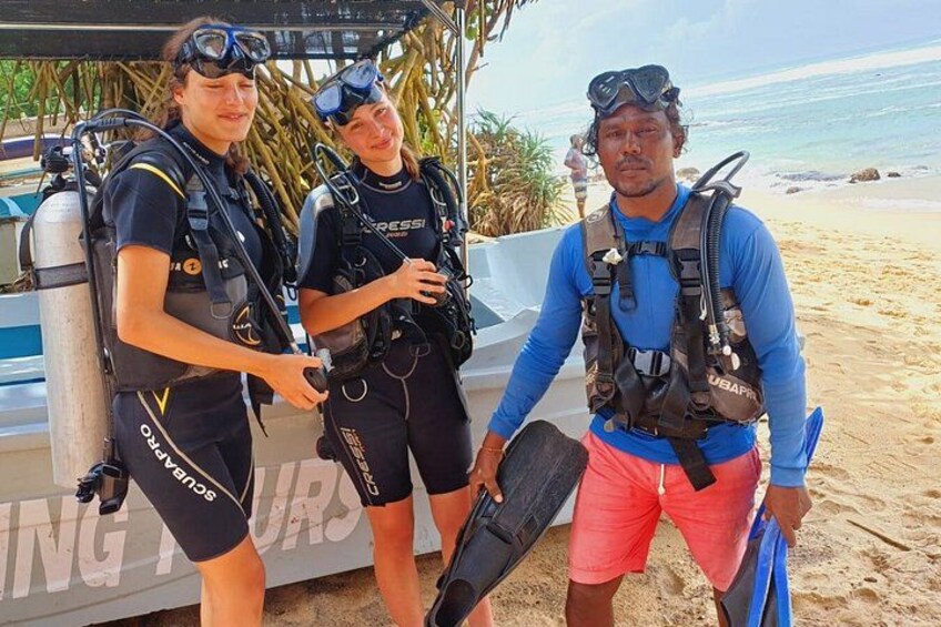 Three people in scuba diving gear standing on a sandy beach with a boat in the background. 1