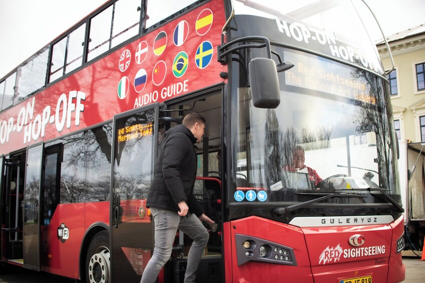 Un autobus à deux étages rouge, portant les inscriptions « montez et descendez à volonté » et « visite guidée audio », est garé et une personne y monte. 1