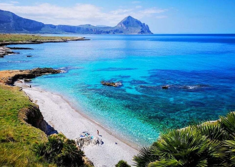 A coastal landscape with clear turquoise waters, a sandy beach, rocky outcrops, and a mountain in the distance. 1
